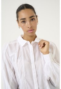 Weiße Bluse mit durchgehender Knopfleiste, mit Puffärmeln und strukturierten Akzenten entlang der Nähte. Weicher, leichter Stoff.