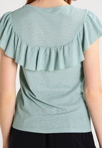 Ljusgrön T-shirt av strukturerad tyg, med en volangdesign på baksidan och korta ärmar. Enkel halsringning och avslappnad passform.