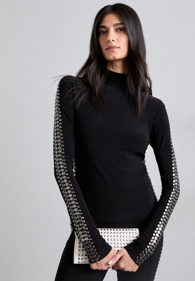 Black long-sleeve top with a high neck, featuring metallic studs along the sleeves. Model holds a studded clutch. Smooth fabric texture.