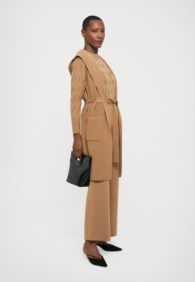 Μπεζ πλεκτή εμφάνιση με υφή από καλωδιώσεις, ζώνη στη μέση και παντελόνι με φαρδιά πόδια, συνδυασμένη με μια μικρή μαύρη τσάντα και μαύρες επίπεδες παπούτσια.