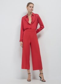 Blusa di raso rosso con bottoni e maniche lunghe abbinata a pantaloni a gamba larga coordinati. Design semplice, texture liscia, tacchi con cinturino alla caviglia.