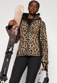 Luipaardprint skijack met een ritssluiting aan de voorkant en zwarte accenten. Het model houdt skies met bindingen vast en draagt zwarte broeken. Glad stoftextuur.