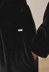 Schwarzer Veloursbademantel mit strukturierter Oberfläche. An der vorderen Tasche befindet sich ein silbernes "DOWNTIME"-Etikett. Lange Ärmel mit lockerer Passform.