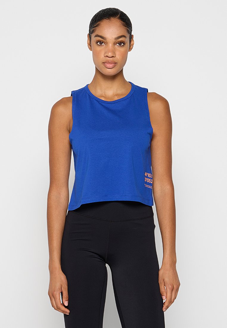 Haut sans manches coupé en coton bleu vif, présentant un col rond et un détail texte orange sur le côté gauche. Porté avec des leggings noirs.