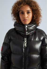 Giacca a vento nera con texture lucida, colletto alto, zip visibile e branding a contrasto. Presenta un accento rosso sulla spalla.
