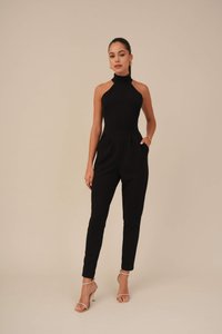 Combinaison sans manches noire avec un col montant, des jambes fuselées et des poches latérales ; fabriquée en tissu doux. Associée à des talons dorés à lanières.