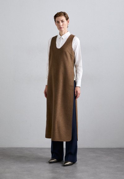 Vrouw in een lange, mouwloze, bruine jurk over een witte blouse, met een marineblauwe wijde broek en metallic puntige schoenen, staande op een grijze vloer.