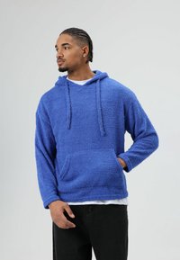 Blauwe fleecehoodie met een gestructureerde afwerking, zak aan de voorkant, capuchon met trekkoord en lange mouwen, gecombineerd met een wit T-shirt eronder.