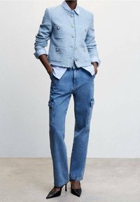 Veste croisée en tweed bleu clair avec boutons argentés, chemise blanche à col, et pantalon cargo en denim bleu avec poches latérales, assortis à des talons noirs.