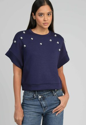 Jeune femme portant un haut à manches courtes bleu marine avec des ornements sphériques argentés et un jean bleu asymétrique à boutons, debout devant un fond uni.