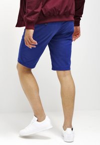 Modré bavlněné kraťasy s hladkým povrchem, ke kolenům, se zadními kapsami a zapínáním na knoflík, v kombinaci s bílými teniskami.