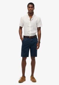 Outfit da uomo con una leggera camicia di lino a maniche corte, pantaloni corti blu navy, cinturino in pelle marrone e scarpe in suede beige con lacci.