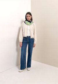 Giacca bomber beige chiaro, t-shirt bianca, jeans larghi blu e sneakers bianche. Sciarpa verde e navy con motivo decorativo.