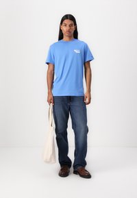 T-shirt en coton bleu clair à manches courtes avec un petit logo blanc sur la poitrine, associé à un jean foncé et un sac fourre-tout beige.