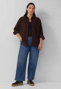 Bruine corduroy button-up shirt over een zwarte top, gecombineerd met lichtblauwe wijde jeans en zwarte enkellaarsjes met hak. Eenvoudige, casual look.