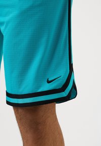Shorts de sport turquoise avec un liseré noir portés sur une jambe, avec un petit logo Nike noir près de l'ourlet sur fond blanc.