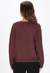 Femeie îmbrăcată cu un pulover tricotat în culoarea vinului, cu modele înċălțate pe mâneci, văzută din spate, purtat cu pantaloni negri.