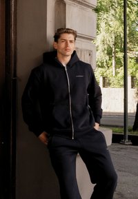 Marineblå zip-up hettegenser laget av mykt stoff, med en frontlomme og snor i hetten. Kombinert med matchende joggebukser.