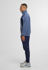 Modrá fleecová bunda na zip s vysokým límcem, námořnické kalhoty a stříbrné běžecké boty. Kontrastní barevné akcenty na bundě a kalhotách.