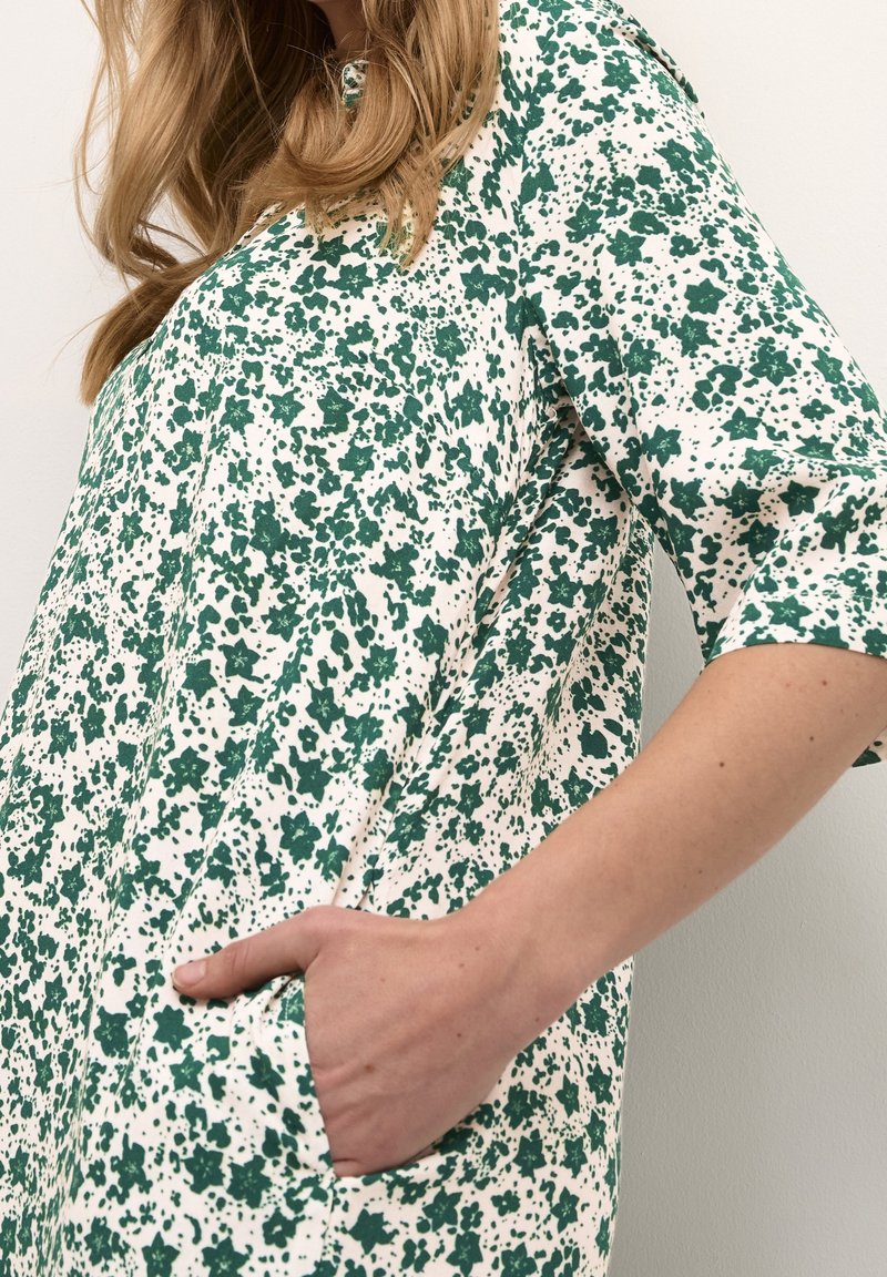 Femme portant une robe blanche avec un motif floral vert, cheveux longs et main dans la poche latérale sur fond uni.