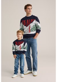 Tumši zilas adītas džemperis ar uzrakstu "LES ALPES" sarkanā krāsā, veidots ar kalnu un koku dizainu. Apvienots ar gaiši zilām džinsām un baltajiem sporta apaviem.