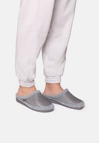 Sive slip-on papuče s tkanim gornjim dijelom, mekanim sivim rubom i izdržljivim potplatom, nosene s svijetlosivim trenirkama.