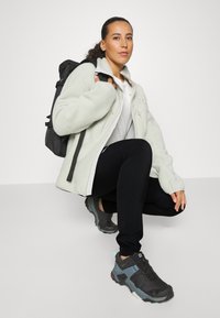 Casaco de fleece verde claro com fecho, leggings pretas e sapatos de caminhada cinza-pretos. Mochila preta pendurada em um ombro.