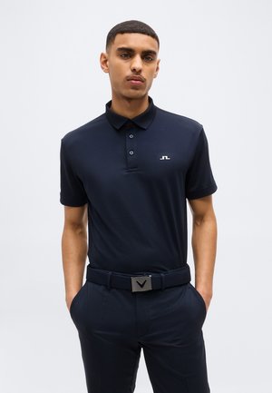 Homme portant un polo bleu marine ajusté à manches courtes et un pantalon bleu marine, les mains dans les poches, debout devant un fond uni.
