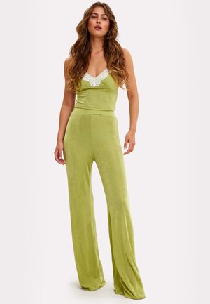 Vrouw met lang golvend haar, gekleed in lichtgroene broek met wijde pijpen en bijpassende mouwloze top met witte kanten rand, staand tegen een witte achtergrond.