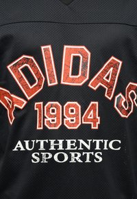 Μαύρο αθλητικό ύφασμα με διάτρητα  μοτίβα και κόκκινο κείμενο "ADIDAS 1994" καθώς και λευκό κείμενο "AUTHENTIC SPORTS" σε έντονα γράμματα.