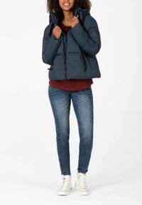 Dunkelblauer Steppjacke mit Kapuze, tailliertem Schnitt, hohem Kragen und Schnürdetails. Getragen über einem bordeauxroten Oberteil und blauen Jeans. Weiße Sneaker.