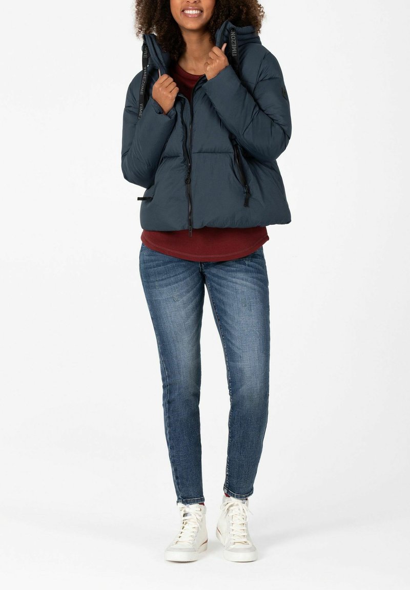 Dunkelblauer Steppjacke mit Kapuze, tailliertem Schnitt, hohem Kragen und Schnürdetails. Getragen über einem bordeauxroten Oberteil und blauen Jeans. Weiße Sneaker.