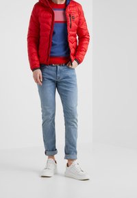 Muž v červené péřové bundě, modro-růžovém pruhovaném svetru, světle modrých džínech s náplety a bílých teniskách stojí proti bílému pozadí.