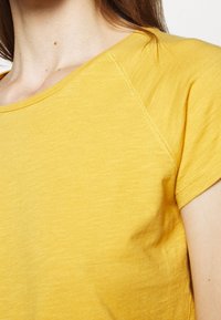 Zbliżenie osoby w żółtej koszuli z krótkimi rękawami, ukazujące dekolt, szew ramienia i górną część ramienia.