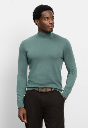 Uomo con maglione a collo alto verde e pantaloni scuri, in piedi con una mano in tasca, che guarda di lato su uno sfondo semplice.