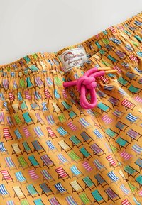 Short de bain avec motif coloré de chaise de plage sur tissu jaune, avec cordon rose noué en boucle et étiquette « Swimwear NEXT ».