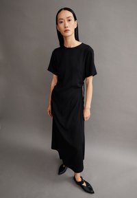 Schwarzes Midi-Kleid mit kurzen Ärmeln, glatter Textur, lässiger Schnitt, seitliches Bindedetail, getragen mit spitzen schwarzen Schuhen vor einem grauen Hintergrund.