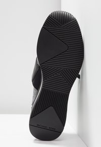 Sole di sneaker nera con battistrada in gomma, caratterizzata da una superficie testurizzata e un logo impresso. Il design include scanalature angolate per una migliore aderenza.
