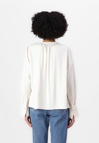 Blouse couleur crème avec manches longues, encolure froncée et coupe décontractée. Le tissu semble lisse avec un léger éclat.