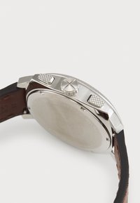 Montre en acier inoxydable avec un bracelet en cuir marron, des accents texturés et une couronne en argent. Le fond du boîtier est poli et rond.