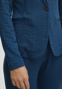 Texturovaná modrá bunda s herringbone vzorem. Má zapínání na jeden knoflík a přední kapsy. Materiál se zdá být tkaný a ušitý na míru.