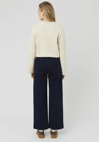 Un pull en maille texturée de couleur crème associé à un pantalon en velours côtelé noir marine à jambes larges, avec des poches arrière et un ourlet droit. Des chaussures marron complètent le look.