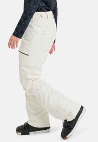 Cremfarbene Hose mit lockerer Passform, ausgestattet mit einer Reißverschlusstasche und Gesäßtaschen, kombiniert mit schwarzen Schuhen und einem gemusterten dunklen Oberteil.