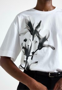 Hvid bomulds T-shirt med et sort blomsterdesign med liljer på forsiden. Løs pasform, korte ærmer, rund hals og glat tekstur.