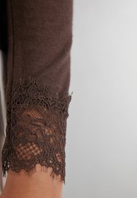 Brun stickad plagg med spetsmanschetter med intrikata blommönster, som kontrasterar med en slät textur och delikata, detaljerade spetskanter.