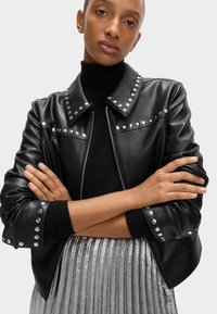 Zwarte leren jas met zilveren studs op de kraag en schouders, voorzien van een rits aan de voorkant en geribbelde mouwen, gedragen over een zwarte coltrui.