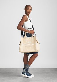 Beżowa torba na ramię z płótna z białym napisem "TORBA PODRÓŻNA" oraz "MARC JACOBS". Posiada mocne uchwyty i kieszenie po bokach. Nosi się z dżinsową spódnicą i niebieskimi sneakersami.