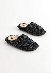 Chaussons noirs tricotés avec un motif texturé, doublure en fourrure beige douce et semelles en caoutchouc sombre. Comprend une petite étiquette marron sur un des chaussons.