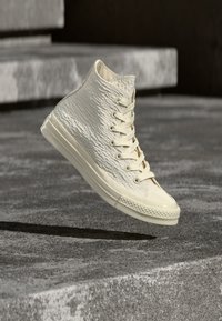 Αθλητικά παπούτσια τύπου high-top σε ανοιχτό μπεζ υλικό με υφή, με κορδόνια και γυαλιστερό καπάκι δαχτύλου, αιωρούμενα πάνω από υφή γκρι επιφάνειας.