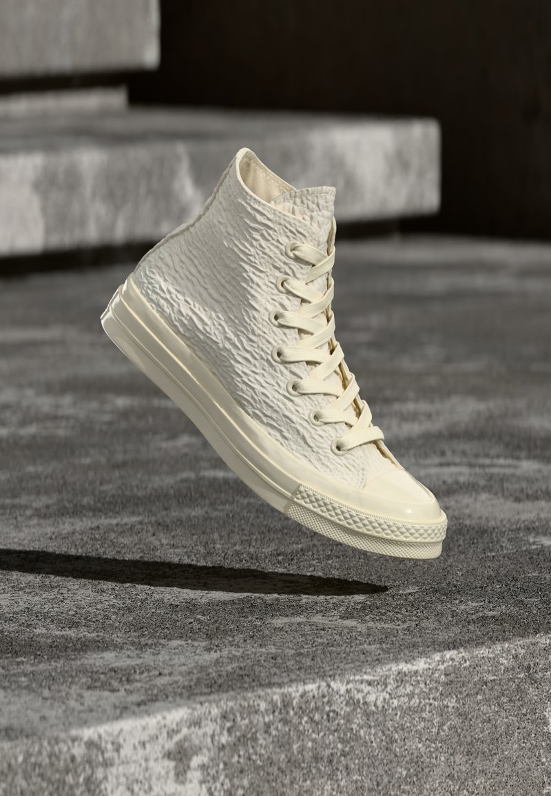 Baskets montantes en matériau texturé blanc cassé avec des détails à lacets et un bout brillant, suspendues au-dessus d'une surface grise texturée.